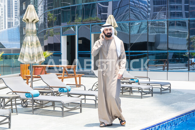 Connecting and communicating with others, using a modern and advanced technological device, answering a mobile phone call, spending enjoyable times exchanging news and conversations, relaxing outdoors, walking along the edge of the water pool, an Emirati Gulf Arab man wearing a kandura and ghutra and taking a stroll.