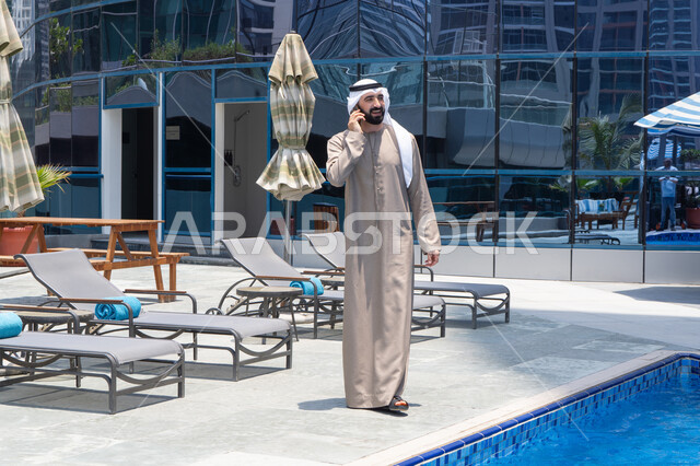 Remote communication, using a modern and advanced technological device, answering a phone call on a mobile phone, spending enjoyable time exchanging news and conversations, relaxing outdoors, walking along the edge of a water feature, an Emirati Gulf Arab man wearing a kandura and ghutra going for a stroll