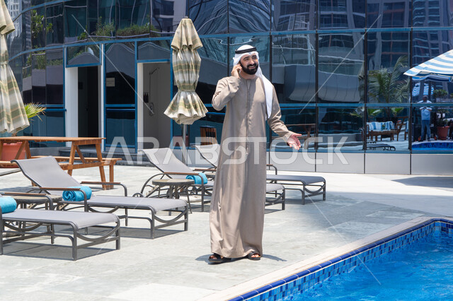 تعابير الاندماج والتركيز بالتحدث عبر الهاتف المحمول، قضاء اوقات ممتعة بتبادل الاخبار والأحاديث،  الإستجمام في الهواء الطلق, المشي على حدود البركة المائية, رجل عربي خليجي اماراتي يرتدي الكندورة والغترة و يقوم بالتنزه