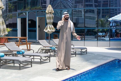 تعابير الاندماج والتركيز بالتحدث عبر الهاتف المحمول، قضاء اوقات ممتعة بتبادل الاخبار والأحاديث،  الإستجمام في الهواء الطلق, المشي على حدود البركة المائية, رجل عربي خليجي اماراتي يرتدي الكندورة والغترة و يقوم بالتنزه