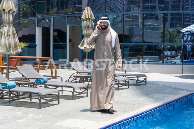 الرد على مكالمة هاتفية عبر الجوال، قضاء اوقات ممتعة بتبادل الاخبار والأحاديث، الإستجمام في الهواء الطلق, المشي على حدود البركة المائية, رجل عربي خليجي اماراتي يرتدي الكندورة والغترة و يقوم بالتنزه