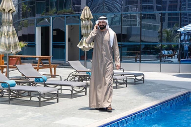 Answering a phone call on a mobile phone, spending enjoyable time exchanging news and conversations, relaxing outdoors, walking along the edge of a pond, an Emirati Gulf Arab man wearing a kandura and ghutra going for a stroll