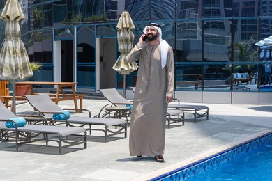 استخدام الأجهزة الحديثة والمتطورة, الرد على مكالمة هاتفية عبر الجوال، قضاء اوقات ممتعة بتبادل الاخبار والأحاديث، الإستجمام في الهواء الطلق, المشي على حدود البركة المائية, رجل عربي خليجي اماراتي يرتدي الكندورة والغترة و يقوم بالتنزه