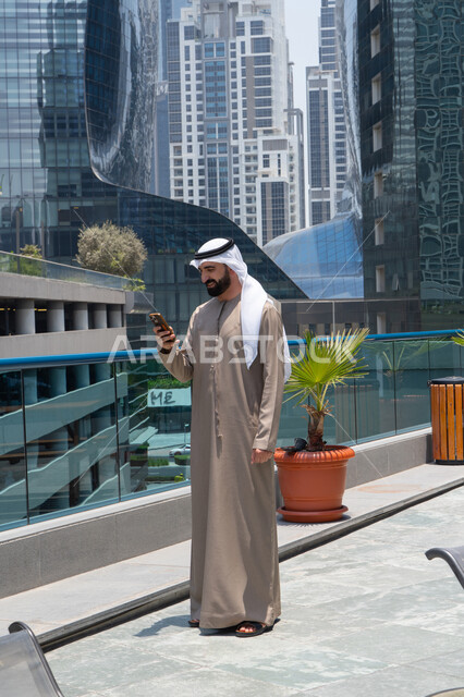 تصفح مواقع التواصل الاجتماعي عبر الانترنت، الإستجمام في الهواء الطلق, المشي على حدود البركة المائية, رجل عربي خليجي اماراتي يرتدي الكندورة والغترة و يقوم بالتنزه و استخدام الموبايل, قضاء الاوقات ممتعة