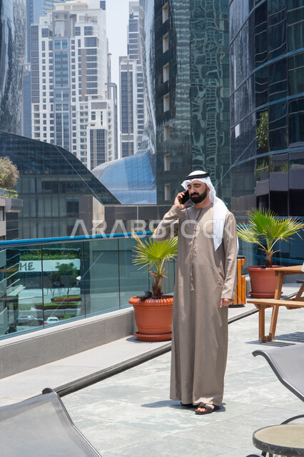 التواصل مع العائلة و الأصدقاء في أوقات الفراغ, مكالمات هاتفية عبر الهاتف المحمول, الدردشة باستخدام الموبايل، الإستجمام في الهواء الطلق, المشي على حدود البركة المائية, رجل عربي خليجي اماراتي يرتدي الكندورة والغترة و يقوم بالتنزه حول الممشى