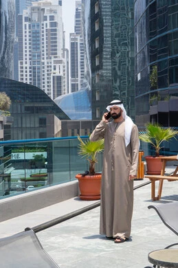 التواصل مع العائلة و الأصدقاء في أوقات الفراغ, مكالمات هاتفية عبر الهاتف المحمول, الدردشة باستخدام الموبايل، الإستجمام في الهواء الطلق, المشي على حدود البركة المائية, رجل عربي خليجي اماراتي يرتدي الكندورة والغترة و يقوم بالتنزه حول الممشى