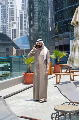 Sharing news with family and friends, chatting on mobile, relaxing outdoors, walking along the edge of the pond, an Emirati Gulf Arab man wearing a kandura and ghutra making a phone call on his mobile phone
