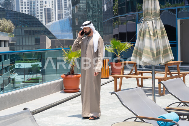 Making a phone call using a mobile phone, exchanging news with family and friends, chatting on a mobile phone, relaxing outdoors, walking along the edge of the water feature, an Emirati Gulf Arab man wearing a kandura and ghutra strolling around the promenade