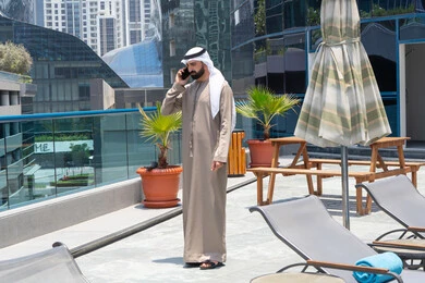 Making a phone call using a mobile phone, exchanging news with family and friends, chatting on a mobile phone, relaxing outdoors, walking along the edge of the water feature, an Emirati Gulf Arab man wearing a kandura and ghutra strolling around the promenade