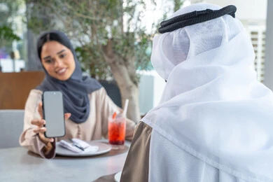 دمج التقنيات الحديثة فى الحياة اليومية ،شرب المشروبات المنعشه، قضاء اوقات ممتعة مع الأصدقاء، بناء وتعزيز العلاقات والصداقات، اجتماع عمل في مكان خارج مقر الشركة, زميلان عمل عربيان خليجيان إماراتيان يجلسان على طاولة في أحد المطاعم الراقية في الإمارات احداهما يعرض شاشة الجوال