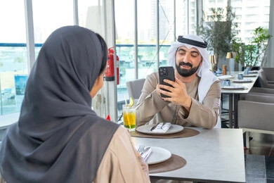 رجل عربي خليجي اماراتي يرتدي الكندورة والغترة يجلس على طاولة في أحد المطاعم الراقية في الإمارات يقوم بالتقاط صورة تذكارية عبر الجوال، توثيق اللحظات السعيدة ، استخدام الهاتف المحمول ، قضاء اوقات ممتعة مع الأصدقاء، بناء وتعزيز العلاقات والصداقات، مناقشة الاعمال وتبادل الافكار، اجتماع عمل في مكان خارج مقر الشركة