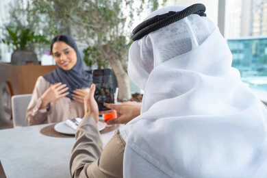 استخدام الهاتف المحمول ، توثيق اللحظات السعيدة ، قضاء اوقات ممتعة مع الأصدقاء، بناء وتعزيز العلاقات والصداقات، مناقشة الاعمال وتبادل الافكار، اجتماع عمل في مكان خارج مقر الشركة, رجل عربي خليجي اماراتي يرتدي الكندورة والغترة يجلس على طاولة في أحد المطاعم الراقية في الإمارات يقوم بالتقاط صورة تذكارية لزميلته عبر الجوال
