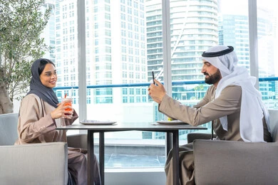 توثيق اللحظات السعيدة ، استخدام الهاتف المحمول ، قضاء اوقات ممتعة مع الأصدقاء، بناء وتعزيز العلاقات والصداقات، مناقشة الاعمال وتبادل الافكار، اجتماع عمل في مكان خارج مقر الشركة, رجل عربي خليجي اماراتي يرتدي الكندورة والغترة يجلس على طاولة في أحد المطاعم الراقية في الإمارات يقوم بالتقاط صورة تذكارية لزميلته عبر الجوال