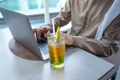 الاستمتاع بالمشروبات الباردة المنعشة ، دمج التقنيات الحديثة فى الحياة اليومية ، قضاء اوقات ممتعة فى الخارج، بناء وتعزيز العلاقات والصداقات، اجتماع عمل في مكان خارج مقر الشركة,صورة مقربة ليد رجل عربي خليجي اماراتي يرتدي الكندورة والغترة يجلس على طاولة في أحد المطاعم الراقية في الإمارات يستخدم الحاسوب المحمول