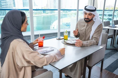 Gestures of happiness and joy, spending enjoyable times with friends, building and strengthening relationships and friendships, discussing business and exchanging ideas, a business meeting in a place outside the company headquarters, two Arab Gulf Emirati colleagues sitting at a table in one of the upscale restaurants in the UAE, exchanging conversations and smiles.