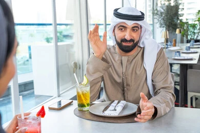 قضاء اوقات ممتعة مع الأصدقاء، إيماءات السعادة والسرور، بناء وتعزيز العلاقات والصداقات، مناقشة الاعمال وتبادل الافكار، اجتماع عمل في مكان خارج مقر الشركة, زميلان عمل عربيان خليجيان إماراتيان يجلسان على طاولة في أحد المطاعم الراقية في الإمارات، يتبادلان الاحاديث والابتسامات