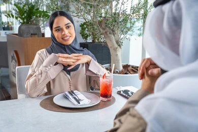إيماءات السعادة والسرور، قضاء اوقات ممتعة مع الأصدقاء، بناء وتعزيز العلاقات والصداقات، مناقشة الاعمال وتبادل الافكار، اجتماع عمل في مكان خارج مقر الشركة, زميلان عمل عربيان خليجيان إماراتيان يجلسان على طاولة في أحد المطاعم الراقية في الإمارات، يتبادلان الاحاديث والابتسامات