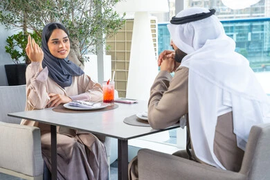 Two Arab Gulf Emirati colleagues are sitting at a table in a high-end restaurant in the UAE, exchanging conversations and smiles, gestures of happiness and joy, enjoying pleasant times with friends, building and strengthening relationships and friendships, discussing business and exchanging ideas, a business meeting in a place outside the company headquarters.