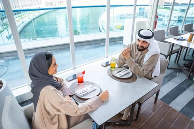 اجتماع عمل في مكان خارج مقر الشركة, إيماءات السعادة والسرور، قضاء اوقات ممتعة مع الأصدقاء، بناء وتعزيز العلاقات والصداقات، مناقشة الاعمال وتبادل الافكار،زميلان عمل عربيان خليجيان إماراتيان يجلسان على طاولة في أحد المطاعم الراقية في الإمارات، يتبادلان الاحاديث والابتسامات