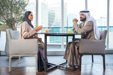 بناء وتعزيز العلاقات والصداقات، إيماءات السعادة والسرور، قضاء اوقات ممتعة مع الأصدقاء، مناقشة الاعمال وتبادل الافكار، اجتماع عمل في مكان خارج مقر الشركة, زميلان عمل عربيان خليجيان إماراتيان يجلسان على طاولة في أحد المطاعم الراقية في الإمارات، يتبادلان الاحاديث والابتسامات