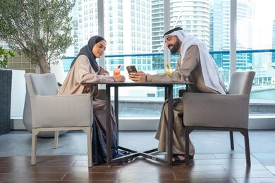 قضاء اوقات ممتعة مع الأصدقاء، توثيق اللحظات السعيدة ، التقاط الصور التذكارية عبر الجوال ، بناء وتعزيز العلاقات والصداقات، مناقشة الاعمال وتبادل الافكار، اجتماع عمل في مكان خارج مقر الشركة, زميلان عمل عربيان خليجيان إماراتيان يجلسان على طاولة في أحد المطاعم الراقية في الإمارات احداهما يستخدم الهاتف المحمول