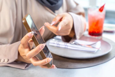 Spending enjoyable times with friends, using mobile phones, building and strengthening relationships and friendships, discussing business and exchanging ideas, a business meeting in a place outside the company headquarters, an Arab Gulf Emirati woman wearing an abaya and hijab sitting at a table in one of the upscale restaurants in the UAE displaying her mobile screen. Spending enjoyable times with friends, using mobile phones, building and strengthening relationships and friendships, discussing business and exchanging ideas, a business meeting in a place outside the company headquarters, an Arab Gulf Emirati woman wearing an abaya and hijab sitting at a table in one of the upscale restaurants in the UAE displaying her mobile screen.