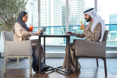 Discussion of business and exchange of ideas, a work meeting in a place outside the company headquarters, two Arab Gulf Emirati colleagues sitting at a table in one of the upscale restaurants in the UAE, one of them using a mobile phone, documenting happy moments, taking commemorative photos via mobile, spending enjoyable times with friends, building and strengthening relationships and friendships.