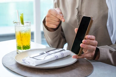 استخدام الهاتف المحمول ، قضاء اوقات ممتعة مع الأصدقاء، بناء وتعزيز العلاقات والصداقات، مناقشة الاعمال وتبادل الافكار، اجتماع عمل في مكان خارج مقر الشركة, رجل عربي خليجي اماراتي يرتدي الكندورة والغترة يجلس على طاولة في أحد المطاعم الراقية في الإمارات يقوم بعرض شاشه الجوال