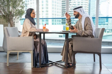 Two Arab Gulf Emirati colleagues are sitting at a table in a high-end restaurant in the UAE. One of them is using a mobile phone to document happy moments, take commemorative photos through the phone, spend enjoyable times with friends, build and strengthen relationships and friendships, discuss business and exchange ideas, and hold a business meeting outside the company premises.