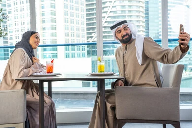 توثيق اللحظات السعيدة ، التقاط الصور التذكارية عبر الجوال ، قضاء اوقات ممتعة مع الأصدقاء، بناء وتعزيز العلاقات والصداقات، مناقشة الاعمال وتبادل الافكار، اجتماع عمل في مكان خارج مقر الشركة, زميلان عمل عربيان خليجيان إماراتيان يجلسان على طاولة في أحد المطاعم الراقية في الإمارات احداهما يستخدم الهاتف المحمول