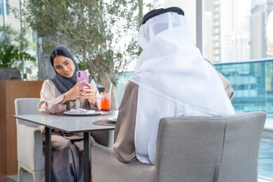 مناقشة الاعمال وتبادل الافكار، اجتماع عمل في مكان خارج مقر الشركة, زميلان عمل عربيان خليجيان إماراتيان يجلسان على طاولة في أحد المطاعم الراقية في الإمارات احداهما يستخدم الهاتف المحمول،توثيق اللحظات السعيدة ، التقاط الصور التذكارية عبر الجوال ، قضاء اوقات ممتعة مع الأصدقاء، بناء وتعزيز العلاقات والصداقات