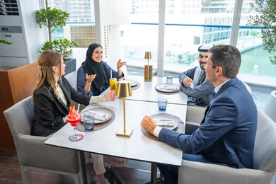 A group of foreign and Arab friends, Emirati Gulf nationals, are sitting at a table in a restaurant in the UAE, sharing enjoyable moments, enjoying cold drinks, having a business meeting outside the company headquarters, and building successful social relationships.