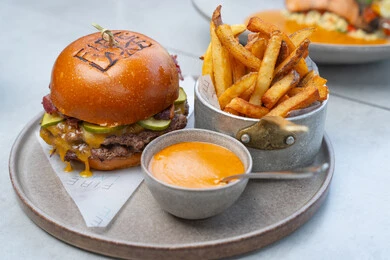 A delicious meal consisting of a premium beef burger topped with melted cheese, pickle slices, and pieces of bacon inside a shiny soft bun, served alongside crispy French fries and a rich-flavored sauce in a side bowl, gourmet meals, Arab Gulf Emirati restaurants, the art of etiquette and presentation.
