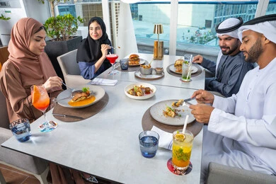 A group of Arab Gulf Emirati friends are sitting at a table in a restaurant in the UAE, a business meeting in a place outside the company headquarters, sharing enjoyable moments, spending fun times with friends, a table filled with various dishes and drinks.