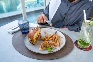 صورة مقربه ليد رجل عربي خليجي اماراتي يستمتع بتناول وجبة شهية مكونة من برجر لحم فاخر مغطى بالجبن الذائب وشرائح المخلل وقطع اللحم المقدد داخل خبز طري لامع، مقدمة بجانب بطاطس مقلية مقرمشة وصوص غني بالنكهة في وعاء جانبي، وجبات الطعام الشهية