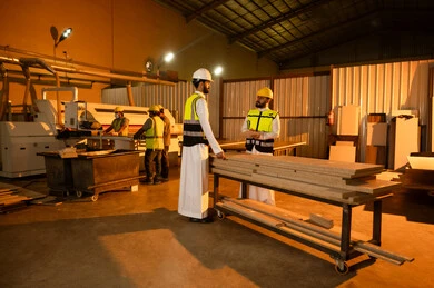 Industrial professions and jobs, exchanging opinions and experiences, carpentry workshops in the Kingdom of Saudi Arabia, wooden furniture manufacturing workshop, a Saudi Gulf Arab engineer wearing a helmet and safety vest standing with a colleague inside the factory discussing the work plan.