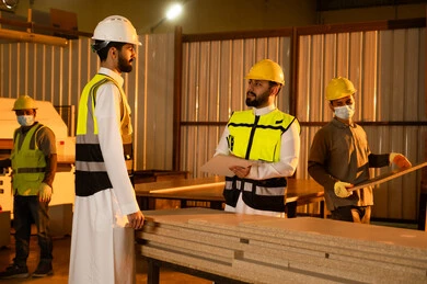 كتابة ملاحظات العمل، جودة الانتاج المحلي الوطني، كتابة و تدوين الملاحظات، مناقشة الخبرات والآراء، صناعة الأثاث المنزلي والمكتبي، ورشات عمل في المملكة العربية السعودية، رجل مهندس عربي خليجي سعودي يرتدي الثوب و خوذة و سترة الحماية يقف برفقة زميلة يقومان بمتابعة سير العمل