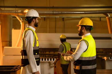 كتابة و تدوين الملاحظات، جودة الانتاج المحلي الوطني، كتابة و تدوين الملاحظات، مناقشة الخبرات والآراء، صناعة الأثاث المنزلي والمكتبي، ورشات عمل في المملكة العربية السعودية، رجل مهندس عربي خليجي سعودي يرتدي الثوب و خوذة و سترة الحماية يقف برفقة زميلة يقومان بمتابعة سير العمل