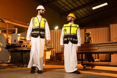 Exchange of opinions and experiences, wooden furniture manufacturing workshop, industrial professions and jobs, monitoring and supervising the workshop machines, carpentry workshops in the Kingdom of Saudi Arabia, two Arab Gulf engineers from Saudi Arabia wearing safety vests and helmets walking inside the factory.