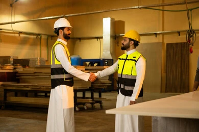 The handshake, greeting, and agreement on tasks, workshops in the Kingdom of Saudi Arabia, the quality of local national production, traditional old crafts and professions, the manufacture of home and office furniture, an Arab Gulf Saudi engineer wearing a white thobe and protective vest is shaking hands with his colleague.