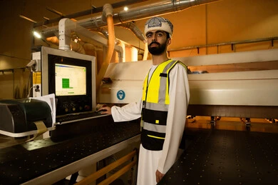 المتابعة والإشراف على آلات المعمل، التحكم بالأجهزة من خلال التكنولوجيا، مصنع تصنيع الأثاث الخشبي، مهن ووظائف صناعية، ورشات النجارة في سلطنه عمان، مهندس عربي خليجي عماني يرتدي العمامة وسترة الحماية يقف داخل المصنع