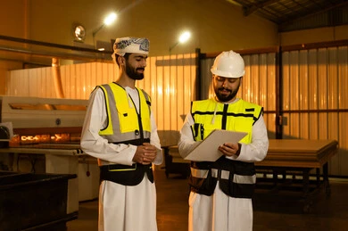 مهن ووظائف عمانية خليجيه، منتج محلي وطني، التحدث في امور العمل، مهندسان عربيان خليجيان عمانيان يرتديان خوذه وستره الحمايه يتناقشان حول خطة العمل ، الحوار والاتفاق في ورشة نجارة الخشب، تسجيل و تدوين الملاحظات