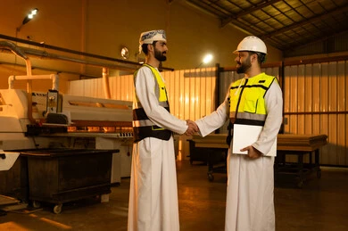 The handshake, greeting, and agreement on tasks, workshops in the Sultanate of Oman, quality of local national production, traditional old crafts and professions, manufacturing of home and office furniture, an Arab Gulf Omani engineer wearing a turban and protective vest is shaking hands with his colleague.