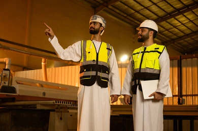 Sharing experiences and opinions, wearing a safety helmet in the factory, pointing to something, local Omani factories and plants, two Arab Gulf engineers from Oman wearing the dishdasha and safety vests are discussing work matters.