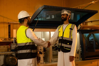Welcoming and greeting and following up on task completion, workshops in the Sultanate of Oman, quality of national local production, traditional old crafts and professions, manufacturing of home and office furniture, an Arab Gulf Omani engineer wearing a turban and protective jacket is shaking hands with his colleague.