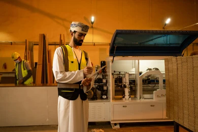 تدوين التحديثات الأخيرة للمشروع، معمل تصنيع الأثاث المنزلي، التطور والتقدم التقني في صناعة الخشب، العمل في القطاع الصناعي، مهندس عربي خليجي عماني يرتدي العمامة و ستره الحماية  يقوم بتدوين و تسجيل الملاحظات، الاشراف على العمال