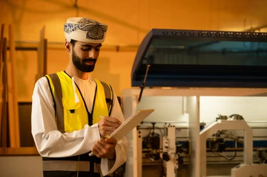 متابعة المستجدات الخاصة بالمشروع، معمل تصنيع الأثاث المنزلي، التطور والتقدم التقني في صناعة الخشب، العمل في القطاع الصناعي، مهندس عربي خليجي عماني يرتدي العمامة و ستره الحماية  يقوم بتدوين و تسجيل الملاحظات، الاشراف على العمال