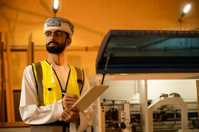 تدوين آخر التطورات في المشروع، معمل تصنيع الأثاث المنزلي، التطور والتقدم التقني في صناعة الخشب، العمل في القطاع الصناعي، مهندس عربي خليجي عماني يرتدي العمامة و ستره الحماية  يقوم بتدوين و تسجيل الملاحظات، الاشراف على العمال