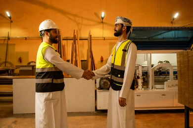 السلام والتحية والاتفاق على انجاز المهام، ورشات عمل في سلطنة عمان, جودة الانتاج المحلي الوطني, مهن وحرف شعبية تقليدية قديمة , صناعة الأثاث المنزلي والمكتبي، رجل مهندس عربي خليجي عماني يرتدي العمامة و ستره الحماية يقوم بمصافحة زميله