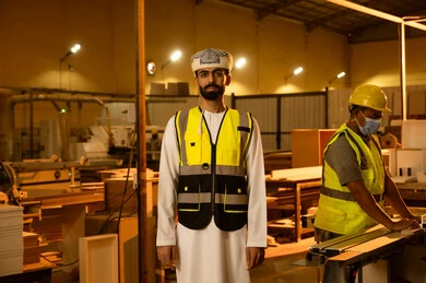 مصنع الأثاث المنزلي، العمل في القطاع الصناعي، التطور والتقدم التقني في صناعة الخشب، عامل يقوم بالعمل على جهاز قص الخشب بالليزر، مهندس عربي خليجي عماني يرتدي العمامة و ستره الحماية يقوم بالاشراف على العمال